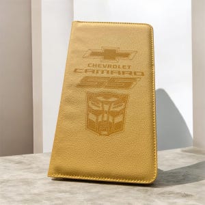 May include: A yellow, textured, rectangular document holder featuring an embossed Chevrolet bowtie logo, the text "CHEVROLET CAMARO 55", and a stylized Autobot face emblem centered on the front surface with visible stitching along the edges.