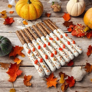 May include: A collection of pretzel rods dipped in white icing and decorated with small orange pumpkin-shaped candies and metallic gold and silver sprinkles, arranged on a wooden surface with scattered autumn leaves and whole pumpkins.