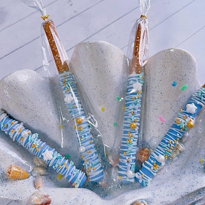 May include: Pretzel rods dipped in light blue chocolate, decorated with white drizzle, star-shaped sprinkles, and gold and white sprinkles. The treats are displayed in a seashell-shaped dish with seashells.