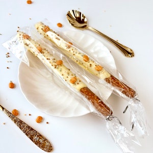 May include: Two pretzel rods dipped in white chocolate, decorated with sprinkles and pumpkin-shaped candies, are wrapped in clear cellophane. The pretzels rest on a white plate, with a gold spoon nearby. Scattered pumpkin candies add a festive touch.