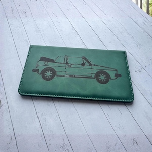 May include: A rectangular green wallet featuring a dark, detailed line art illustration of a convertible car centered on the front surface. The wallet has visible stitching along the edges and a smooth, matte finish across the entire exterior.