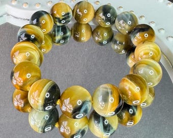May include: A bracelet made of polished, round tiger eye beads. The beads are primarily golden-brown with dark brown and black stripes. The bracelet is strung on a gold-colored cord and is displayed on a white, decorative surface.