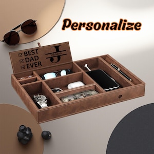 May include: A brown leather valet tray with multiple compartments, holding a watch, phone, glasses, and other accessories. The lid is personalized with the text "Best Dad Ever" and a name. Sunglasses are placed on the top left. The word "Personalize" is written on the top right.