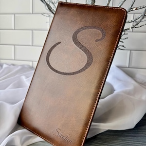 May include: A brown leather journal with the letter "S" engraved on the front. The name "Stephen" is also engraved at the bottom. The journal is rectangular and has a smooth texture. The background includes a white brick wall and silver decorative branches.