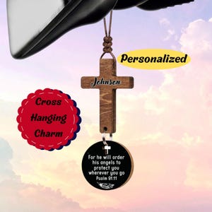 May include: A wooden cross-shaped hanging charm with the name "Johnson" engraved on it. A circular charm with the text "For he will order his angels to protect you wherever you go Psalm 91:11" is attached. The words "Cross Hanging Charm" and "Personalized" are also visible.