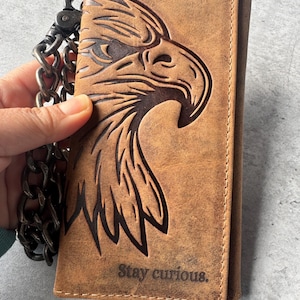 May include: Brown leather wallet featuring an embossed eagle design and the text "Stay curious." The wallet is attached to a chain. The leather has a distressed appearance.