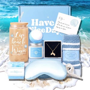 May include: A beach-themed gift box containing a glass tumbler, a gold-coloured necklace with a tail pendant, a candle, bath bomb, eye mask, striped socks, and soap. The box has the text "Have a Nice Day".