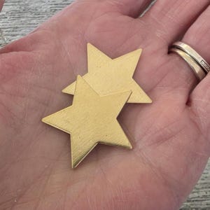 May include: Two gold-colored star-shaped ornaments. The stars are made of a brushed metal material. The stars are overlapping each other. The image is a close-up shot of the stars.