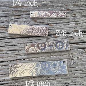 Puede incluir: Cuatro dijes rectangulares de metal plateado con patrones florales decorativos. Los dijes están etiquetados con sus tamaños: 0,6 cm, 1 cm y 1,3 cm. Los dijes están sobre una superficie de madera envejecida.