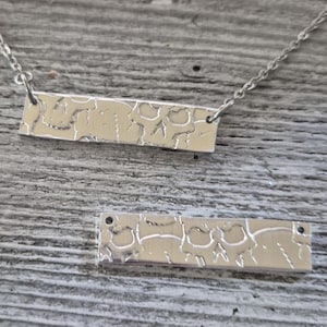 May include: A silver bar necklace with a textured surface and a matching pendant. The necklace features a delicate chain and a rectangular pendant with a unique, patterned design. The pendant is approximately 1.5 inches long.