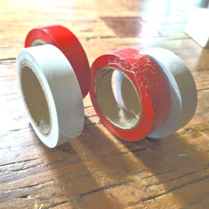 May include: Close-up of four rolls of tape. Two rolls are white, and two are red. The tape rolls are on a wooden surface. The rolls are different sizes and are partially unrolled.