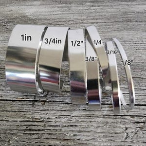 May include: A collection of silver metal strips of varying widths, labeled with measurements in inches: 1in, 3/4in, 1/2in, 1/4in, 3/16in, and 1/8in. The strips are arranged on a weathered, light-colored wooden surface.