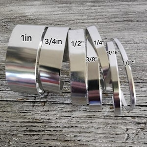 May include: A collection of silver metal strips of varying widths, labeled with their measurements in inches: 1in, 3/4in, 1/2in, 1/4in, 3/16in, and 1/8in. The strips are arranged on a weathered, light-colored wooden surface.