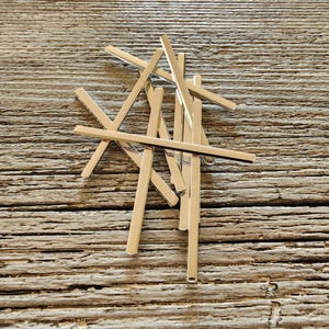 May include: A collection of shiny, silver-colored metal bars arranged on a weathered, wooden surface. The bars are rectangular and appear to be jewelry components or craft supplies. The wood grain adds a rustic contrast to the polished metal.