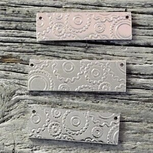 Puede incluir: Tres dijes rectangulares de metal plateado con un diseño en relieve detallado de círculos y líneas onduladas. Cada dije tiene dos pequeños agujeros para sujetar a joyas u otras manualidades. Los dijes son de color plateado y descansan sobre una superficie de madera envejecida.