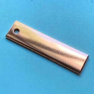 May include: A rectangular, copper-colored metal bar with a hole drilled near one end. The bar has a polished, reflective surface and is set against a solid, light blue background. The bar appears to be approximately 4 inches long.