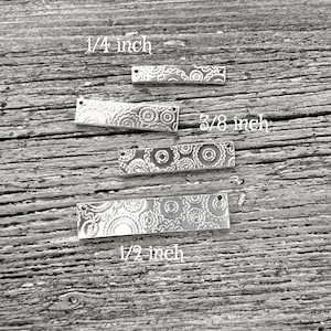 Puede incluir: Cuatro dijes rectangulares de metal plateado con patrones florales decorativos. Los dijes miden 0,6 cm, 1 cm y 1,3 cm de largo. Cada dije tiene un pequeño agujero en cada extremo. Los dijes se muestran sobre una superficie de madera envejecida.