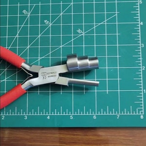 May include: Red handled pliers with a silver metal head and a coiled metal attachment. The pliers are on a green cutting mat with white markings.