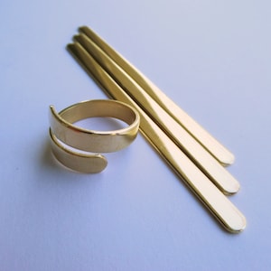 May include: A gold-colored adjustable ring with a spiral design, next to three thin, rectangular gold bars. The jewelry pieces are displayed on a white surface. The ring is a wrap-around style.