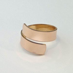 May include: A gold-colored ring with a wrap-around design. The ring has a smooth, matte finish and a simple, elegant style. The band wraps around the finger, creating a modern and minimalist look. The ring is likely made of gold or a gold-toned metal.