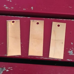May include: Three rectangular copper tags with a hole at the top for hanging. The tags have a polished, reflective surface and are arranged on a red surface. The tags are a warm, metallic color.