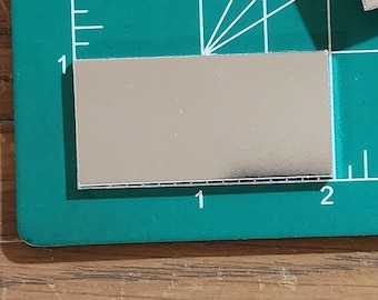 Könnte beinhalten: Ein rechteckiges silbernes Metallblech auf einer grünen Schneidematte mit weißen Markierungen. Das Blech ist 2,5 cm x 5 cm groß.