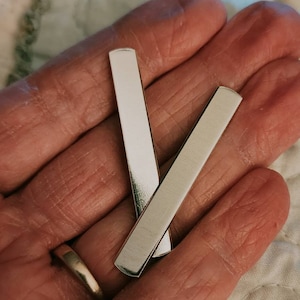 May include: Two silver metal bars, approximately 2 inches long, are held in a hand. The bars are smooth and have rounded edges.
