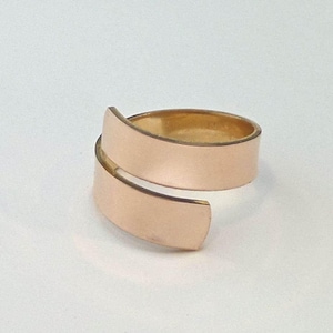 May include: A gold-colored, wrap-style ring. The ring features a smooth, brushed metal finish and a minimalist design. The band wraps around the finger, creating a modern and elegant look. The ring is set against a white background.