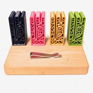Pode incluir: Quatro pequenos blocos geométricos impressos em 3D nas cores preto, rosa, dourado e verde são exibidos em um suporte de madeira. Um clipe de dinheiro de metal prateado também está no suporte.