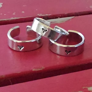 May include: Three silver-colored adjustable rings with a hammered texture and a small heart stamped on each. The rings are displayed on a red surface, creating a contrast with the metallic finish of the jewelry.