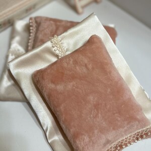 May include: A close-up of two decorative pillows. One pillow is a soft, peach-colored faux fur, and the other is a cream-colored satin. The satin pillow has a delicate lace trim with small pearl-like beads. The pillows are layered.
