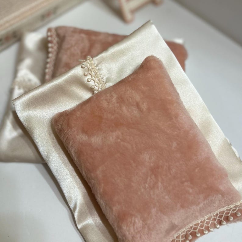 May include: A close-up of two decorative pillows. One pillow is a soft, peach-colored faux fur, and the other is a cream-colored satin. The satin pillow has a delicate lace trim with small pearl-like beads. The pillows are layered.