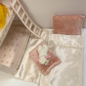 May include: A miniature dollhouse bedroom set with a white ladder, pink and white bedding, and a plush white bunny toy with pink ears. The set includes a pink pillow and a satin-like blanket.