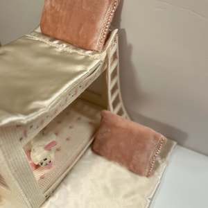 May include: A miniature bunk bed with a cream-colored frame and bedding. The top bunk has a peach-colored pillow with lace trim. The bottom bunk has a peach pillow and a small stuffed lamb. The bedding is cream-colored satin.