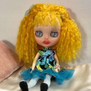 May include: A Blythe doll with bright yellow curly hair and large blue eyes. The doll wears a black dress with a blue and green butterfly pattern, a blue tulle skirt, and striped socks. A pink pillow is in the background.