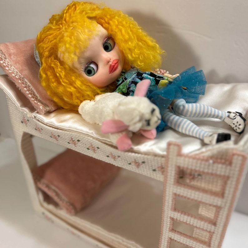 May include: A doll with yellow curly hair and green eyes rests on a miniature bunk bed. The doll wears a blue tutu and striped stockings, and holds a white stuffed bunny with pink accents. The bed has pink pillows and floral trim.