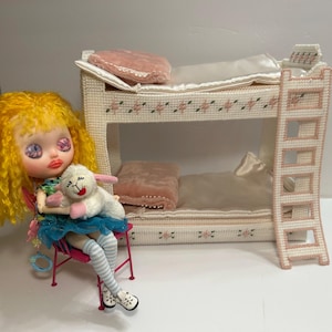 May include: A doll with yellow hair and a blue skirt sits on a pink chair, holding a white lamb plush toy. A miniature, cream-colored bunk bed with pink accents and a ladder is in the background. The bed has pink pillows and floral details.
