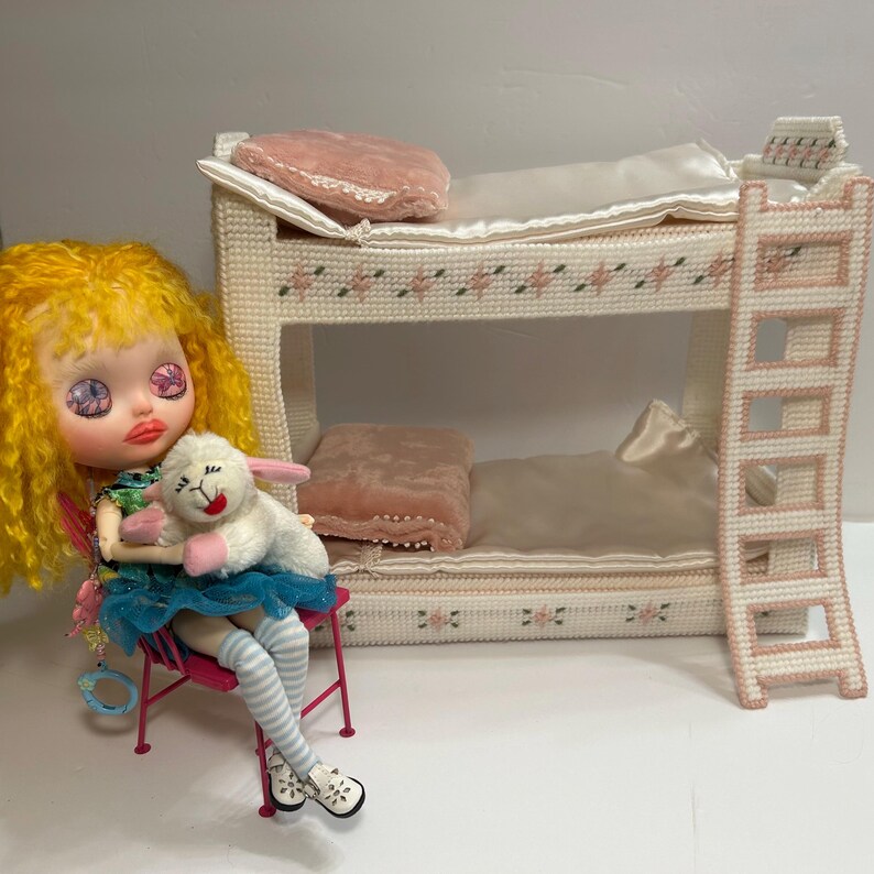 May include: A doll with yellow hair and a blue skirt sits on a pink chair, holding a white lamb plush toy. A miniature, cream-colored bunk bed with pink accents and a ladder is in the background. The bed has pink pillows and floral details.