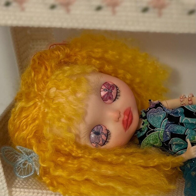May include: A doll with bright yellow hair and butterfly-themed eyes. The doll has a fair complexion, red lips, and is wearing a patterned top. A light blue butterfly decoration is visible near the doll's head. The doll is lying on a cream-colored surface.