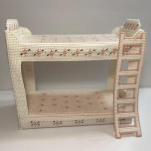 May include: A miniature, handcrafted bunk bed in a cream color, adorned with pink floral and green leaf accents. The bed features a ladder for access to the top bunk. The design is a cross-stitch pattern, giving it a textured appearance.