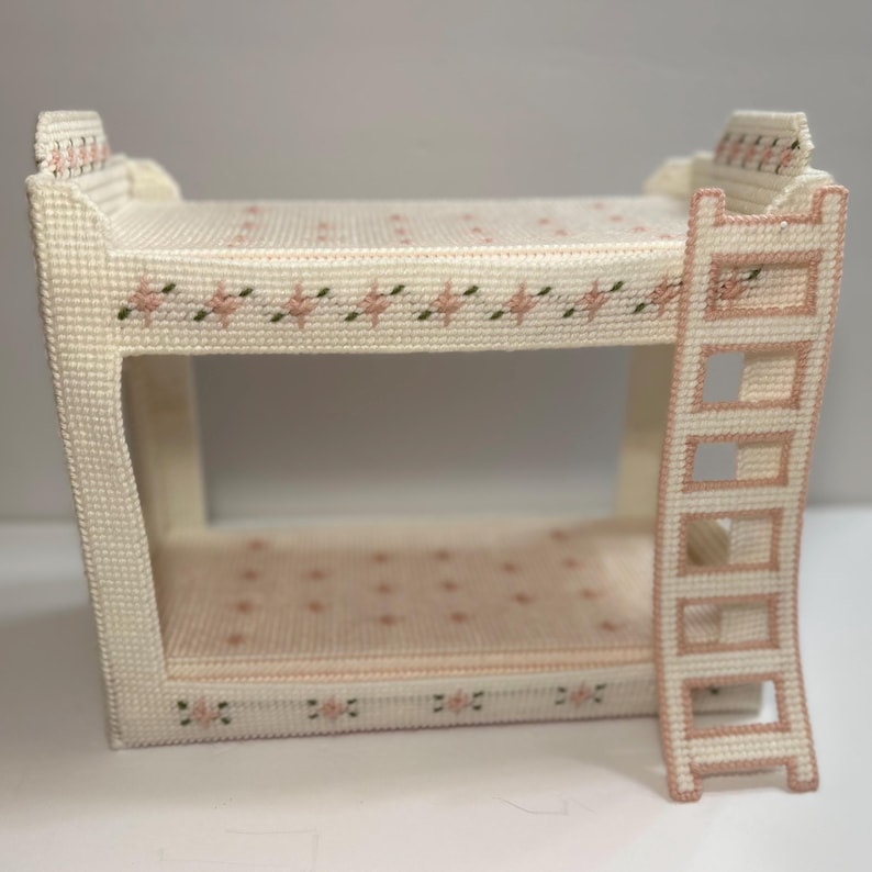 May include: A miniature, handcrafted bunk bed in a cream color, adorned with pink floral and green leaf accents. The bed features a ladder for access to the top bunk. The design is a cross-stitch pattern, giving it a textured appearance.