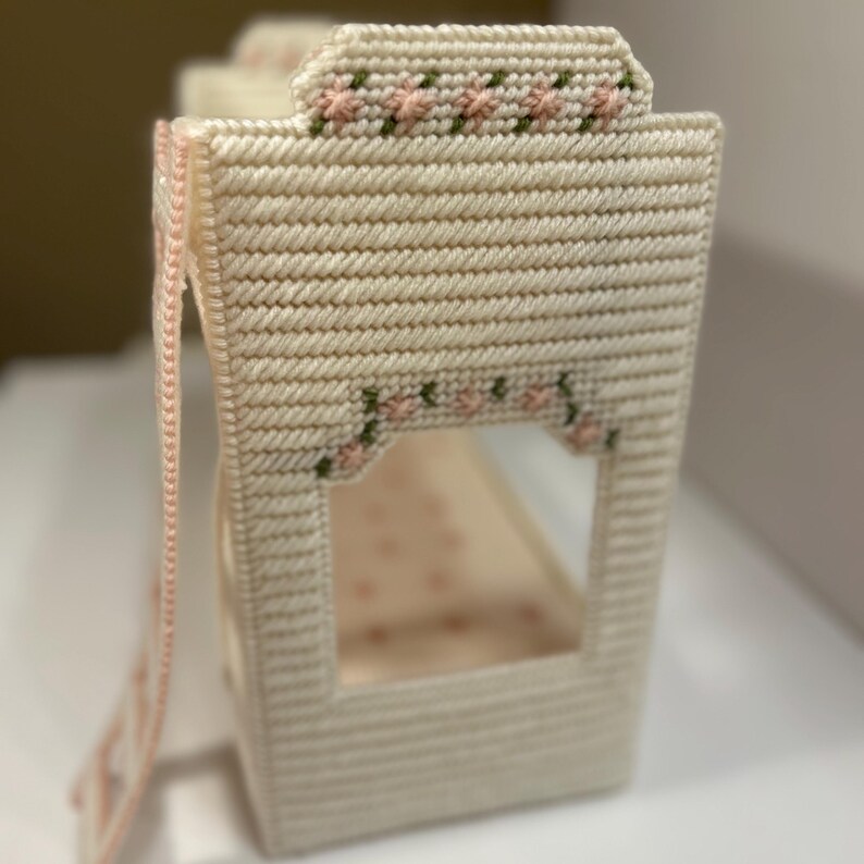 May include: A miniature, off-white, needlepoint bunk bed with pink and green floral accents. The bed features a ladder and decorative floral patterns along the top and window openings. The interior of the bed is a light pink color.