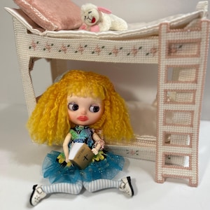 May include: A doll with yellow hair and a blue dress holds a book labeled "Holy Bible." A miniature bunk bed with a pink pillow and a stuffed lamb are in the background. The bed frame and ladder are white with pink accents.