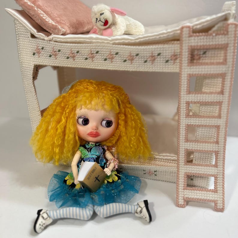 May include: A doll with yellow hair and a blue dress holds a book labeled "Holy Bible." A miniature bunk bed with a pink pillow and a stuffed lamb are in the background. The bed frame and ladder are white with pink accents.
