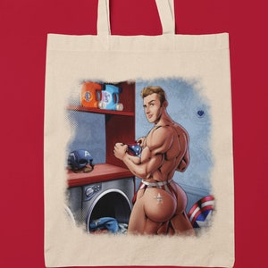 Pode incluir: Uma bolsa tote de lona branca com uma ilustração colorida de um homem musculoso em uma lavanderia com tema vermelho, branco e azul. O homem está segurando uma xícara de café e usando uma roupa íntima com padrão vermelho, branco e azul. A imagem inclui uma máquina de lavar roupa, garrafas de detergente para roupa e uma máscara de super-herói.