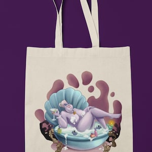 May include: Off-white tote bag featuring a cartoon illustration of a purple character relaxing in a seashell bathtub, holding a cocktail. The design includes a purple and blue color scheme, with the words "TOTE BAG" at the top and bottom.