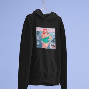 Puede incluir: Sudadera negra con un estampado gr&aacute;fico de una portada de revista que presenta una sirena con cabello rojo y un traje de ba&ntilde;o verde. La portada de la revista se titula "Mermpolitan" y presenta el texto "Total Chic" y "The Male Model & The Prince".