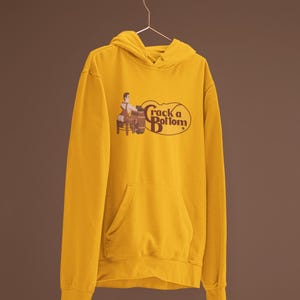 May include: Mustard yellow hoodie with a front pocket and a drawstring hood. The graphic on the front says "Crack a Bottom" with an illustration of a person and a barrel. The hoodie is hanging on a metal rod.