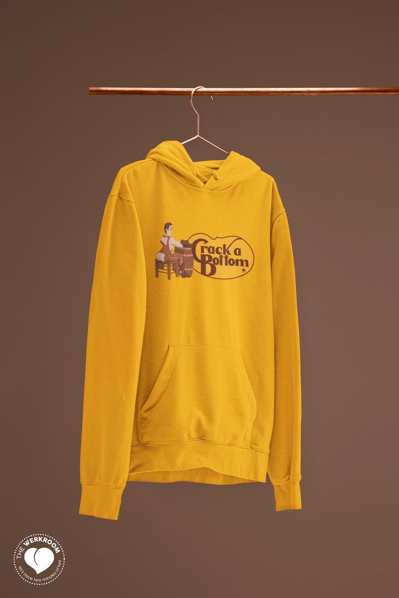 May include: Mustard yellow hoodie with a front pocket and a drawstring hood. The graphic on the front says "Crack a Bottom" with an illustration of a person and a barrel. The hoodie is hanging on a metal rod.