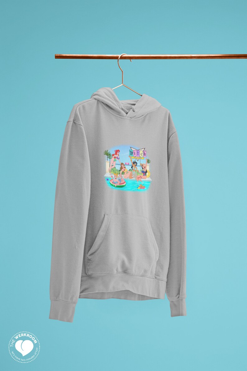 Może przedstawiać: Jasnoszara bluza z kapturem z przednią kieszenią i kapturem ze sznurkiem. Grafika z przodu przedstawia scenę przy basenie z kobietami, palmami i napisem "Bitch". Bluza z kapturem wisi na złotym wieszaku na niebieskim tle.